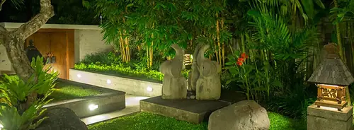 A garden lit at night: two stone figures on a paved plinth, an uplit low wall, bamboo and a stone lantern